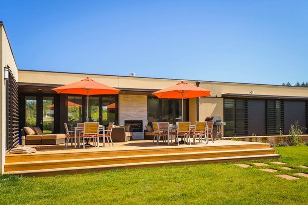 Single-storey contemporary home viewed from the garden, with a broad timber deck and stepped timber platform opening to an outdoor dining and lounge area. Full-height glazing and dark horizontal screening frame the facade, while bright orange umbrellas provide a strong outdoor accent. A manicured lawn and stepping stones sit in the foreground under clear daylight.