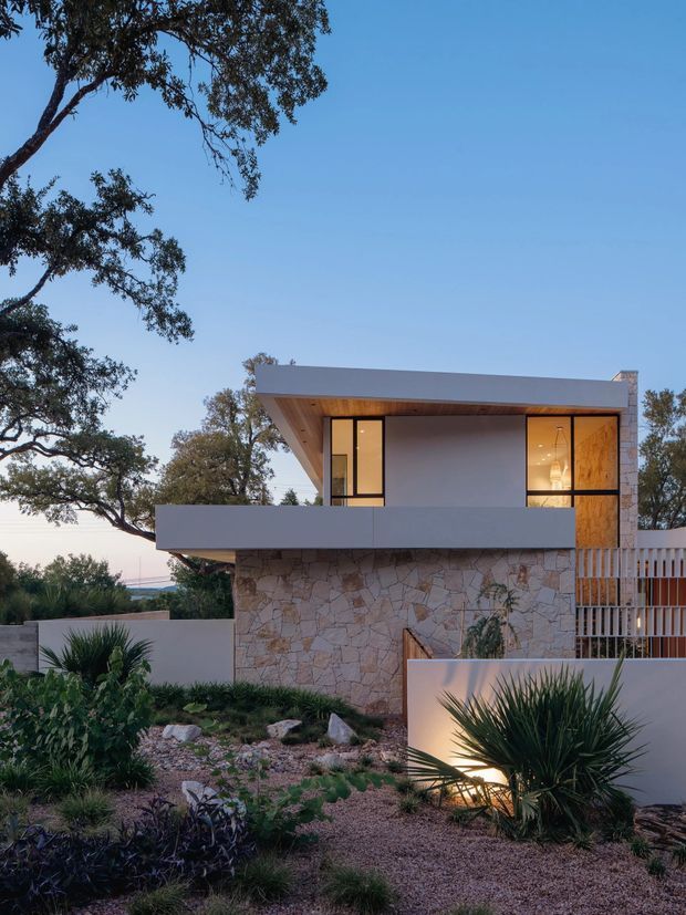 Exterior view of a contemporary two-storey house set within a landscaped garden. The home features a strong flat roof with deep cantilevered eaves, expansive dark-framed glazing, a warm timber-lined soffit, white rendered walls, and a lower facade clad in irregular natural stone. Gravel planting beds, sculptural grasses, succulents, and mature trees surround the building, with a glimpse of distant landscape beyond.