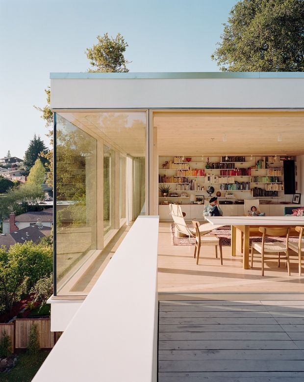 A modern, light-filled open-plan living and dining space viewed from an elevated exterior deck. Floor-to-ceiling glazing wraps the room, with pale timber flooring and ceiling, a full wall of built-in bookshelves, a timber dining table and chairs, and a small seating area. The home overlooks a leafy suburban neighbourhood and is framed by a clean, rectilinear facade with a flat roof.