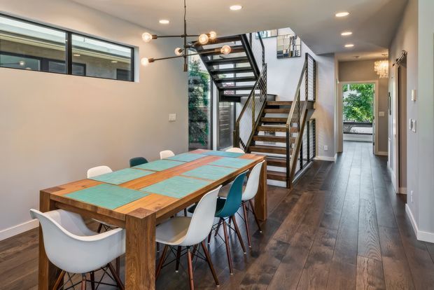 A contemporary open-plan dining and circulation area with a large rectangular timber dining table surrounded by mixed modern chairs. A dramatic open-riser staircase with black metal balustrades rises beside full-height glazing, while a hallway extends toward a bright exterior opening. Recessed ceiling lights and a sculptural globe pendant illuminate dark timber flooring, light painted walls, and the colourful tiled tabletop.