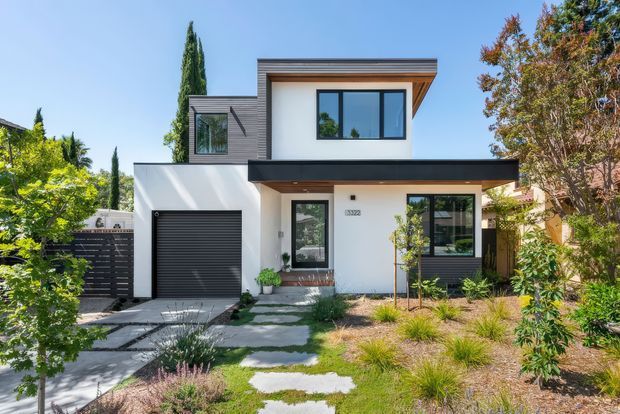 Front exterior view of a contemporary two-storey home with a flat roof, white rendered walls, dark horizontal cladding accents, black-framed glazing, an integrated single garage, and a landscaped front path with drought-tolerant planting.