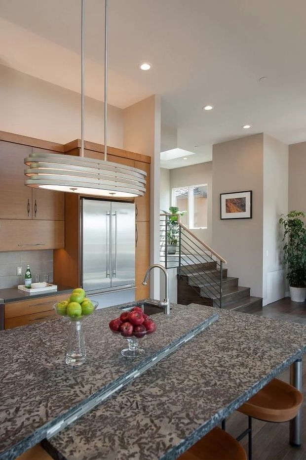 A bright open-plan kitchen featuring a large patterned stone island with an undermount sink and curved chrome faucet, warm timber cabinetry, a stainless-steel double refrigerator, and a layered horizontal pendant light. A staircase and adjacent living circulation area are visible beyond the kitchen. The space has recessed ceiling lighting, neutral walls, and a contemporary elevated interior.