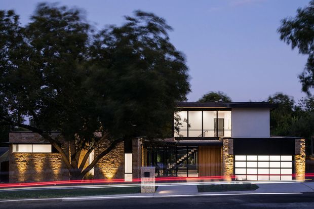 Twilight exterior view of a contemporary multi-level residence with a flat roof, strong horizontal composition, expansive upper-level glazing, dark cladding, warm timber entry detailing, and illuminated textured stone walls. A mature tree partially screens the facade, while light trails from passing traffic appear along the street in the foreground.