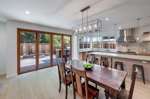 Open-plan dining and kitchen interior with a large dark timber dining table and matching chairs in the foreground. The kitchen includes a long island with bar stools, pale cabinetry, a patterned tile splashback, timber benchtops, and a stainless-steel rangehood. Full-height timber-framed glazed doors open to an outdoor deck, while a linear pendant chandelier hangs above the dining table. The room is bright, contemporary, and warm, with pale timber flooring and white walls.
