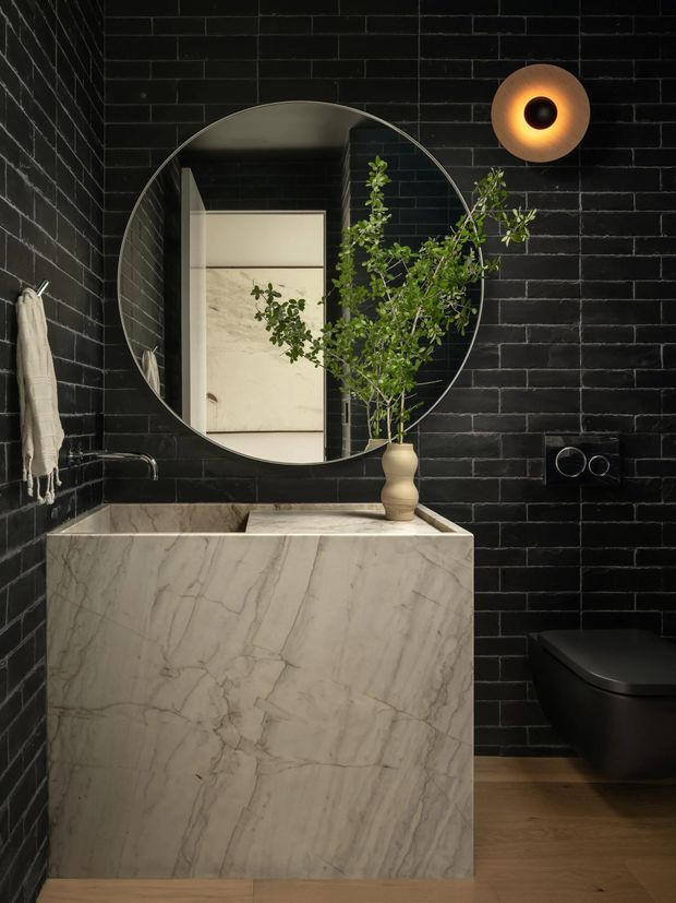 A moody, compact bathroom with glossy black brick wall tiles, a sculptural pale marble floating vanity and integrated basin, a large round mirror, black wall-mounted fixtures, and a concealed black toilet. Warm accent lighting and timber flooring soften the dark, dramatic palette. A doorway and adjacent space are visible in the mirror reflection.