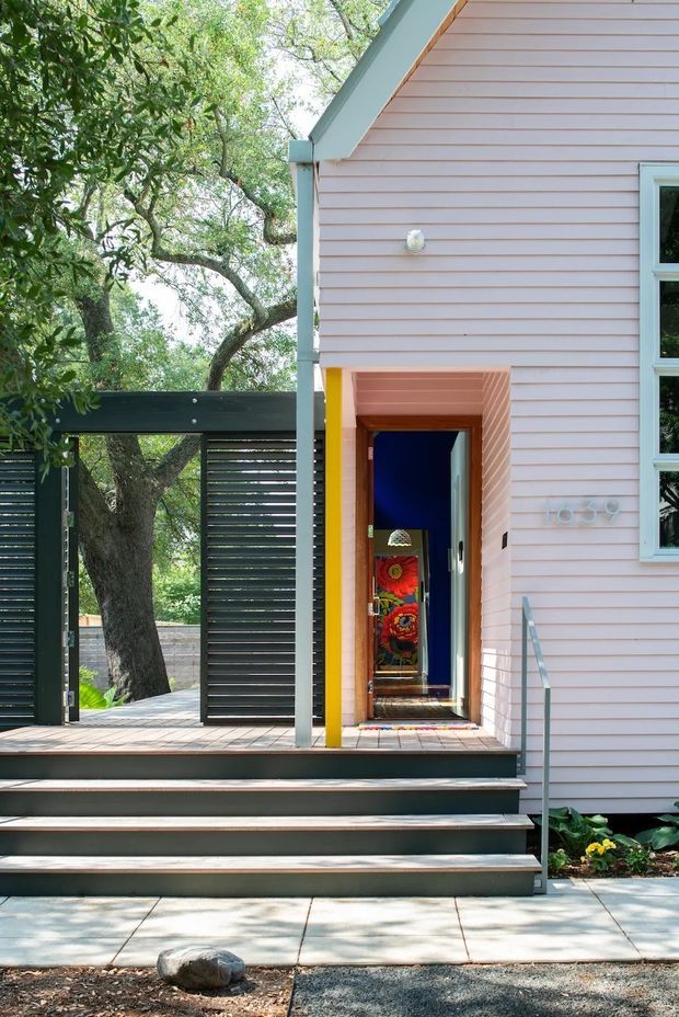 Exterior view of a colourful residential entry with pale pink horizontal weatherboard cladding, white trim, a steep gabled roof edge, and a vivid yellow-framed recessed doorway. Broad timber steps lead up to a raised deck, bordered by dark horizontal slatted screening and a simple metal handrail. Mature trees surround the entry, and a brightly decorated interior is visible through the open door.