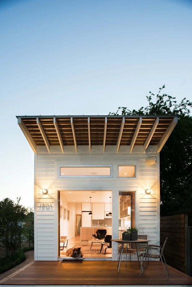 A compact, single-storey backyard dwelling or studio with a white horizontal weatherboard facade, broad exposed timber roof overhang, and large glazed opening onto a timber deck. The illuminated interior appears to contain a small kitchen and seating area, while outdoor dining furniture occupies the deck. The design has a simple contemporary cabin-like form with warm timber detailing and evening lighting.
