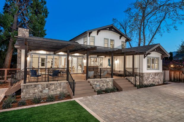 Exterior view of a large, multi-section residential house at dusk, featuring white vertical siding, dark trim, multiple gabled roof forms, broad timber pergolas, covered outdoor sitting areas, stone-clad low walls, timber decking, black metal railings, and a paved driveway or terrace. Warm interior lighting glows through the windows and doors, with landscaped lawn and mature trees surrounding the property.