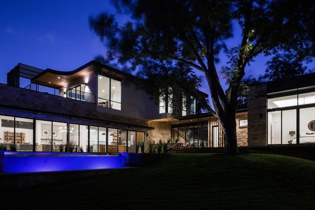 A contemporary multi-wing residence photographed at dusk, with strong horizontal volumes, extensive floor-to-ceiling glazing, a rooftop terrace with dark metal balustrades, and warm interior lighting. A blue-lit swimming pool occupies the foreground, while a large established tree rises through the landscaped lawn. Stone-textured and dark rendered exterior surfaces contrast with the illuminated interiors.