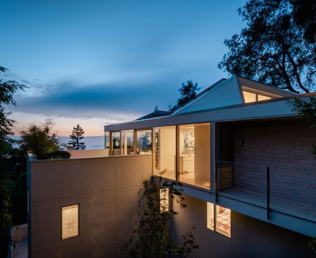 Twilight exterior view of a contemporary multi-level residence with a low-pitched roof, broad floor-to-ceiling glazing, warm interior illumination, rendered walls, and a timber-clad garage or entry volume. The home is set among mature trees with a distant body of water or horizon visible beyond the property.