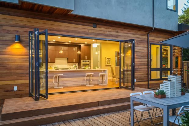 A contemporary timber-clad home opening onto a timber deck, with large bi-fold doors revealing a warmly lit kitchen and island. An outdoor dining setting with an umbrella occupies the foreground, while dark upper-level cladding and recessed lighting add contrast.
