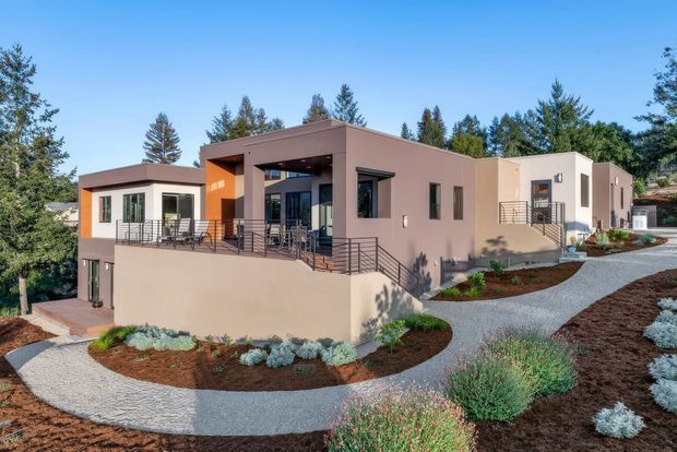 Exterior view of a contemporary, low-profile multi-wing home set within a landscaped garden. The building has smooth rendered walls in muted taupe, cream and orange accents, broad flat roof planes, large dark-framed windows, covered outdoor terraces, and metal balustrades. Curving gravel pathways and drought-tolerant planting surround the raised house.