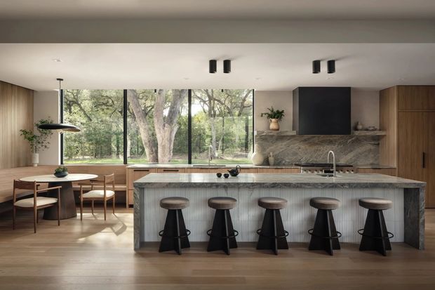A spacious contemporary kitchen and dining area with a large stone island, integrated cooktop, undermount sink and five dark bar stools. The island has a vertically fluted front, while warm timber cabinetry and timber flooring frame a marble- or stone-look splashback and benchtop. Floor-to-ceiling black-framed glazing opens views to mature trees, and a round dining table sits beside the window. Minimal black ceiling lights and a dark rangehood add contrast.