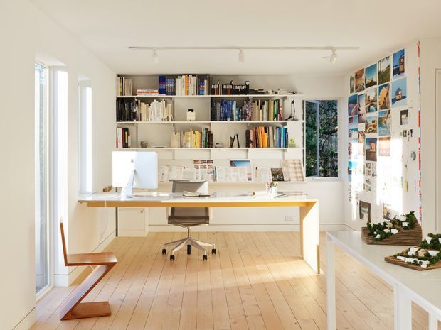 Bright, airy home office or studio with white built-in shelving filled with books and magazines, a large white desk with computer, an office chair, timber flooring, extensive glazing, and a wall displaying photographs and creative materials. The space has a clean, functional, gallery-like character with abundant natural light.