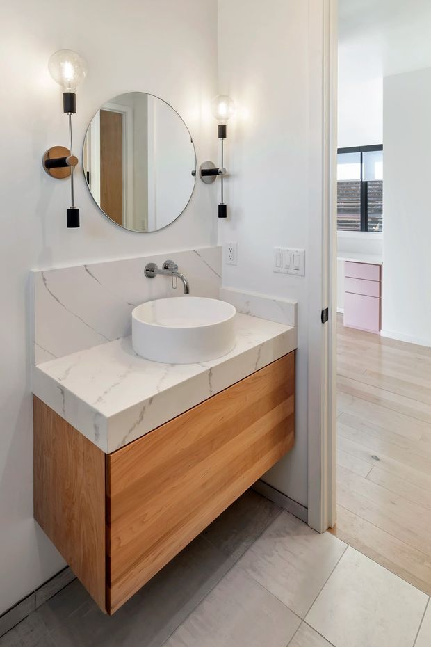 Bright contemporary bathroom vanity with a floating natural timber drawer unit, white veined stone benchtop and integrated backsplash, round vessel basin, wall-mounted chrome tapware, circular mirror, and paired black-and-brass wall sconces. A bedroom or adjoining room with pale timber flooring and pink cabinetry is visible through the open doorway.