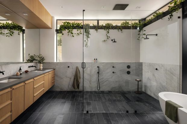 Spacious contemporary bathroom with a large walk-in shower enclosed by frameless glass, dual black shower fixtures, a freestanding bathtub partially visible at right, and a timber vanity with dark benchtop at left. Grey marble-look wall cladding contrasts with dark charcoal floor tiles, while hanging greenery and clerestory glazing add a natural, airy feel. A partial mirror reflects the vanity area.
