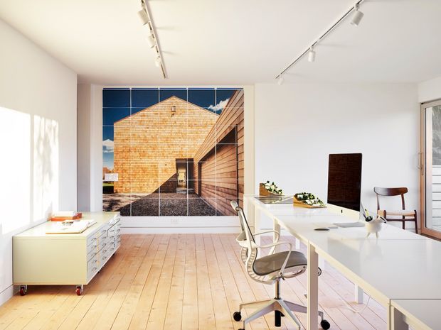 Bright, spacious contemporary office or studio with white walls, pale timber plank flooring, long white worktables, wheeled office chairs, track lighting, and a large architectural mural or photographic wall panel featuring a timber-clad building. A low mobile drawer unit and small dining-style chair add functional furnishings. Sunlight creates strong geometric shadows across the room.