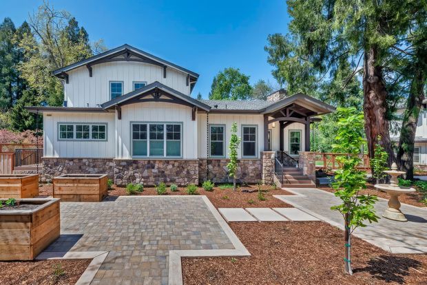 Front exterior view of a suburban two-storey home with a light painted vertical-panel facade, gabled roof forms, stone-clad lower walls, dark timber accents, and a covered entry porch. The landscaped front yard includes raised timber garden beds, paved paths and a broad interlocking-paver driveway or forecourt.