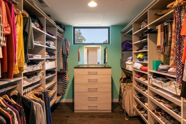 A compact walk-in wardrobe with open built-in shelving and hanging rails on both sides, densely organised with clothing, shoes, bags and folded items. A central light-wood drawer unit with a mirror sits beneath a small high window. The room has teal-green painted walls, recessed ceiling lighting and dark timber flooring.