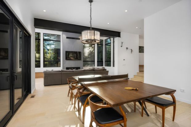 A bright, open-plan dining and living area with a large rectangular timber dining table and curved wooden chairs in the foreground. Behind it is a grey sectional sofa facing a fireplace and wall-mounted artwork, with tall black-framed windows overlooking greenery. A contemporary pendant light hangs above the dining table, while stairs lead upward at the right. The space has pale timber flooring, white walls, dark built-in cabinetry, and exposed dark ceiling beams.