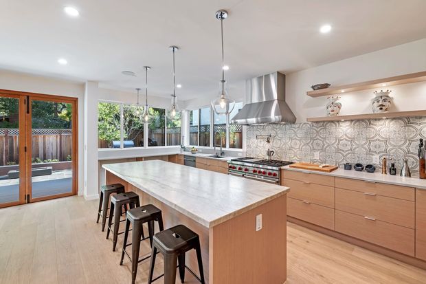 Bright, spacious contemporary kitchen with a large central island, integrated seating for four, light timber cabinetry, pale stone benchtops, patterned tile splashback, stainless-steel rangehood and appliances, pendant lights, and timber-framed glazed doors opening to an outdoor deck.