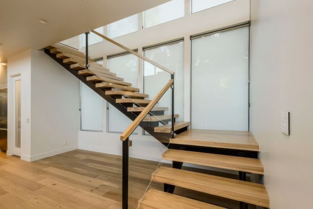 Bright contemporary stairway with floating timber treads, black structural supports, and clear glass balustrades. The staircase rises beside tall frosted-glazed windows, with light timber flooring and white walls creating an airy open interior.