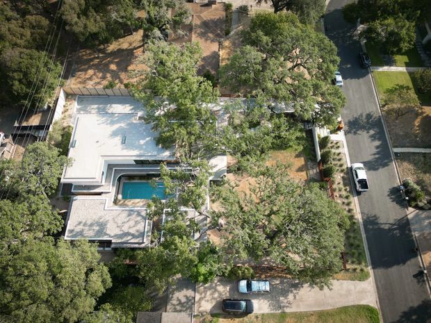 Aerial view of a leafy suburban residential property, showing a flat-roofed modern home partially concealed by mature trees. The property includes a compact swimming pool, paved outdoor areas, landscaped garden spaces, driveway access, and surrounding streets with parked vehicles.