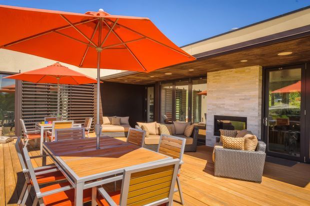 A sunny outdoor entertaining terrace with timber decking, a large dining table and chairs beneath bright orange umbrellas, and a covered lounge area adjoining a modern single-storey residence. The facade combines dark-framed glazing, a warm timber soffit, and a pale stacked-stone fireplace wall.