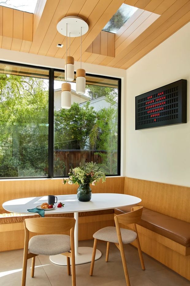 Bright, warm dining nook with a white oval table surrounded by timber dining chairs and a built-in timber banquette. The room features extensive honey-toned timber wall and ceiling panelling, large black-framed corner glazing, sculptural pendant lights, and a black geometric wall artwork. A skylight brings additional natural light into the space, with leafy outdoor greenery visible through the windows.