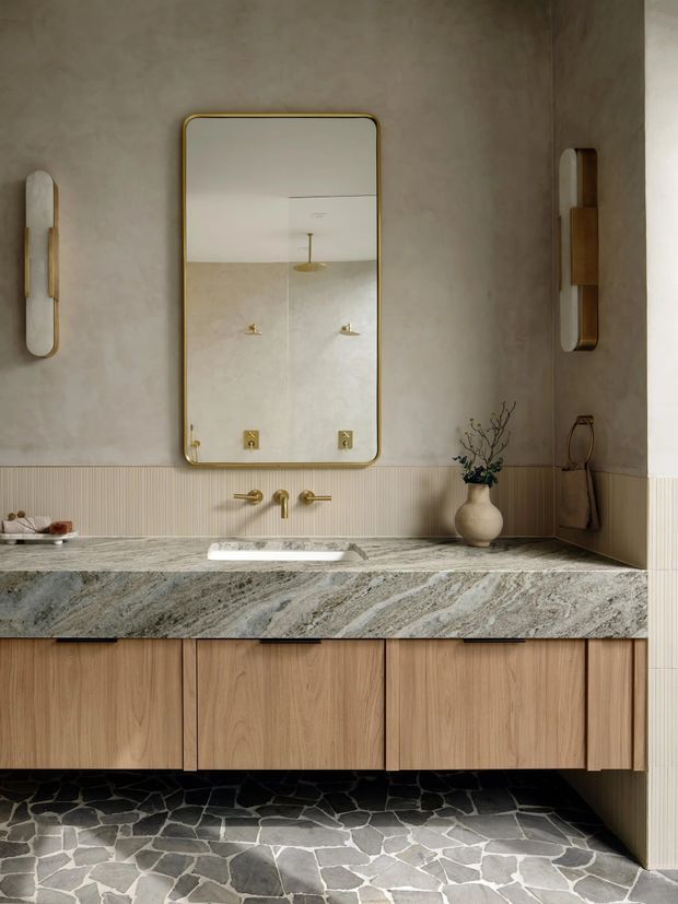A refined contemporary bathroom vanity viewed straight on. The room features a large rounded-rectangle mirror with a brass-toned frame, warm plaster walls, vertically ribbed wall panelling, brass wall sconces and tapware, a veined natural-stone benchtop with an integrated white basin, and floating light timber cabinetry. The floor is finished in irregular dark and light stone pieces, creating a warm, earthy, textured palette.