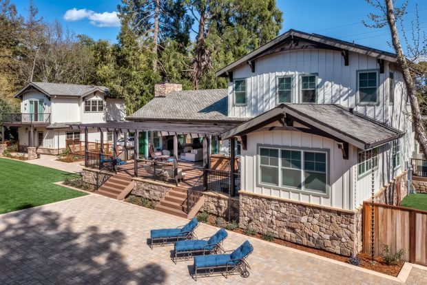 Exterior view of a large multi-level residential property surrounded by mature trees. The building features white vertical cladding, dark grey pitched shingle roofs with exposed timber gable details, stone veneer along the lower walls, and multiple covered outdoor verandah or deck areas with timber stairs and railings. A paved courtyard with blue lounge chairs occupies the foreground, with artificial turf and fencing visible at the sides.