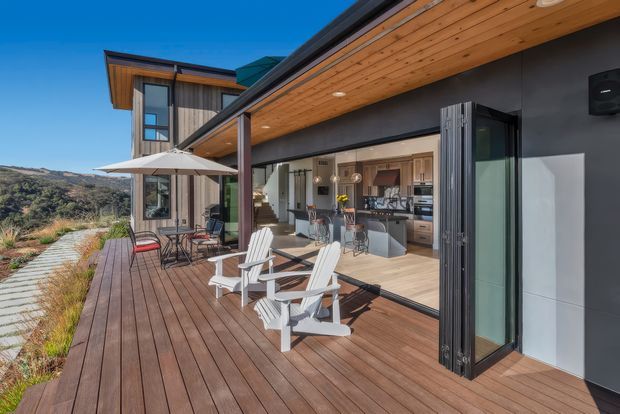 A contemporary outdoor deck adjoining a modern home, with broad timber decking, a covered timber soffit, dark exterior walls, and large bi-fold or sliding doors opening into a bright kitchen and dining area. Outdoor lounge chairs, a small dining setting, and an umbrella occupy the deck, with landscaped planting and a rugged hillside visible beyond.