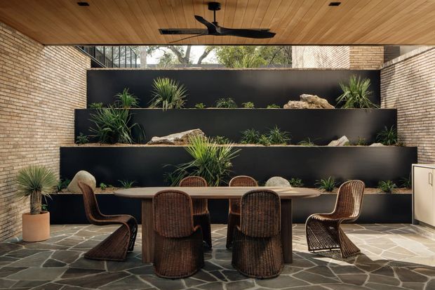A stylish dining area in a partially enclosed courtyard, featuring a long timber dining table surrounded by woven rattan chairs. The space has a warm timber-lined ceiling with a black ceiling fan, exposed light brick walls, dark tiered planter walls with greenery and rocks, and an irregular dark stone-paved floor. A high glazed opening brings in natural light and views of trees.