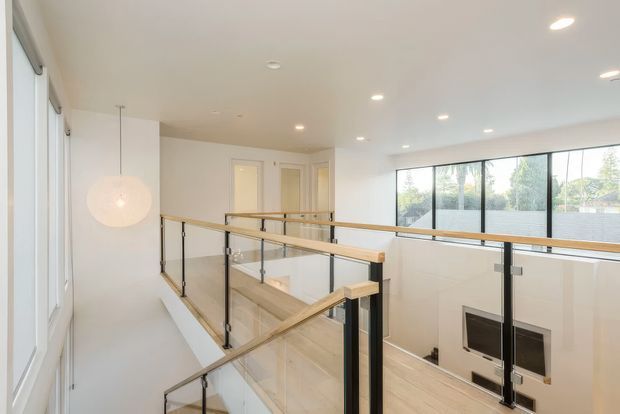 Bright contemporary upstairs landing overlooking a lower-level living area, with pale timber handrails, black metal posts, clear glass balustrades, white walls and ceiling, recessed downlights, and extensive floor-to-ceiling glazing. A fireplace is visible below, while the space is defined primarily by the elevated circulation walkway and stair guardrail.