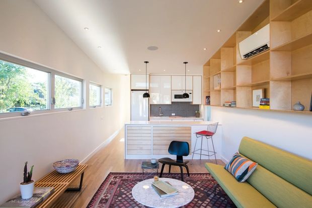 Bright open-plan mid-century-inspired living room and kitchen with a long built-in timber shelving wall, compact kitchen island, timber flooring, large windows, and a colourful green sofa. The space includes a patterned area rug, coffee table, pendant lights, and a restrained contemporary fit-out with warm natural materials.