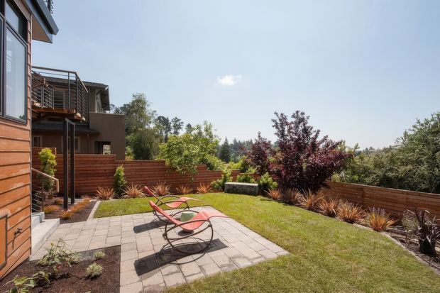 A landscaped residential backyard viewed from beside a modern timber-clad home. The garden features a paved patio with two red reclining lounge chairs, a central lawn, raised timber privacy fencing, ornamental grasses, shrubs, and mature trees. A neighbouring or adjoining modern structure with balconies is visible at the left. The scene is sunny and contemporary, with elevated views toward a leafy suburban landscape.