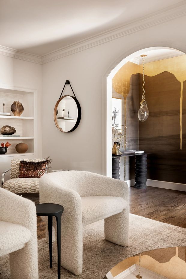 Elegant, warmly lit sitting room with sculptural cream boucle armchairs, a small black side table, a patterned upholstered accent chair, built-in display shelving, and a round wall mirror. An arched opening leads to a dramatic adjoining space featuring a large abstract dark brown and gold wall artwork, a black pedestal table, and a pendant light. The room has white walls with ornate crown moulding, dark timber flooring, and a neutral textured rug.