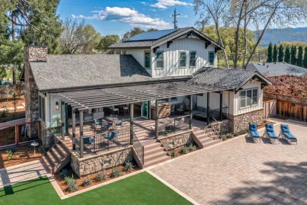 Exterior view of a multi-level suburban house with a grey shingle roof, light vertical siding, stone-clad lower walls, covered outdoor veranda and pergola, paved patio, artificial lawn, and outdoor seating. A mountain ridge and mature trees are visible in the background.