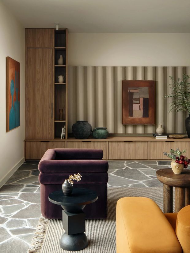 A warm, refined living room featuring a long built-in timber media and storage wall with vertical fluted detailing, open display shelving, and low cabinetry. A deep plum velvet armchair faces a small dark round side table, while a mustard-yellow sofa occupies the foreground. The room includes abstract artwork, ceramic vessels, a textured neutral rug, and a striking irregular grey stone floor with light grout lines. The palette is earthy and sophisticated, with strong mid-century influences.
