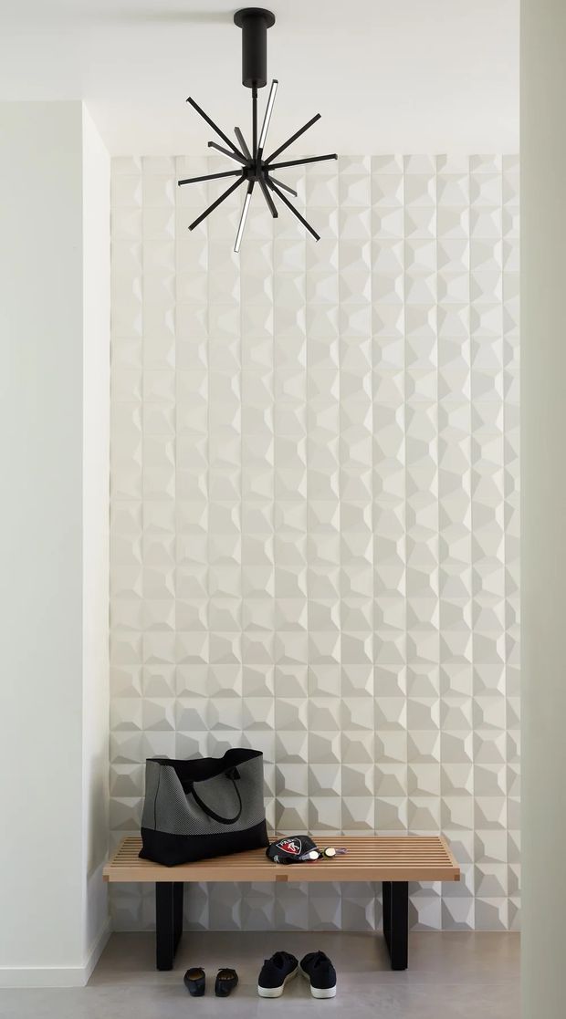 A tall, narrow entryway with a full-height white geometric three-dimensional tiled feature wall. A black starburst pendant light hangs above a simple timber slatted bench with black metal legs, while shoes and a bag are placed below and on the bench. The space is bright and minimalist with pale walls and flooring.