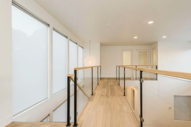Bright modern mezzanine landing or upper-level hallway with pale timber flooring, frameless glass balustrades, slim black metal posts, and warm timber handrails. Tall frosted windows line the left side, while recessed ceiling lights and white walls create an airy, minimalist interior. The lower level is visible through the balustrade opening.