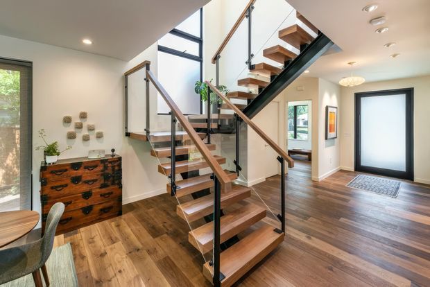 Bright contemporary residential entryway and stair hall featuring an open-riser timber staircase with black metal stringers, timber handrails, and clear glass balustrades. The space has warm timber flooring, white walls, recessed ceiling lights, a large frosted-glass entry door, and a timber chest of drawers with small decorative wall objects. A dining area is partially visible at left and a sitting or living area is visible beyond the stairs.