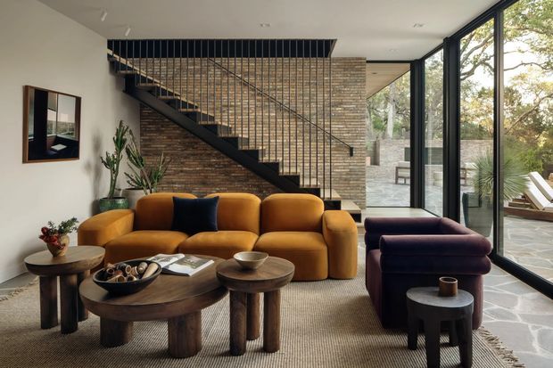 A sophisticated contemporary living room with a mustard modular sofa, dark purple lounge seating, and sculptural timber coffee tables arranged on a neutral textured rug. A black steel stair with slender balustrade rises against a warm exposed brick feature wall, while full-height black-framed glazing opens to a landscaped outdoor area. The room has a warm, refined, earthy character with strong natural light and indoor planting.