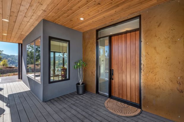 Covered exterior entry porch with a large vertical timber front door, adjacent glazing, dark grey wall volume, timber-lined ceiling, and dark decking. The entry looks over a landscaped, hilly setting and connects visually to the interior through the glazed side panels.