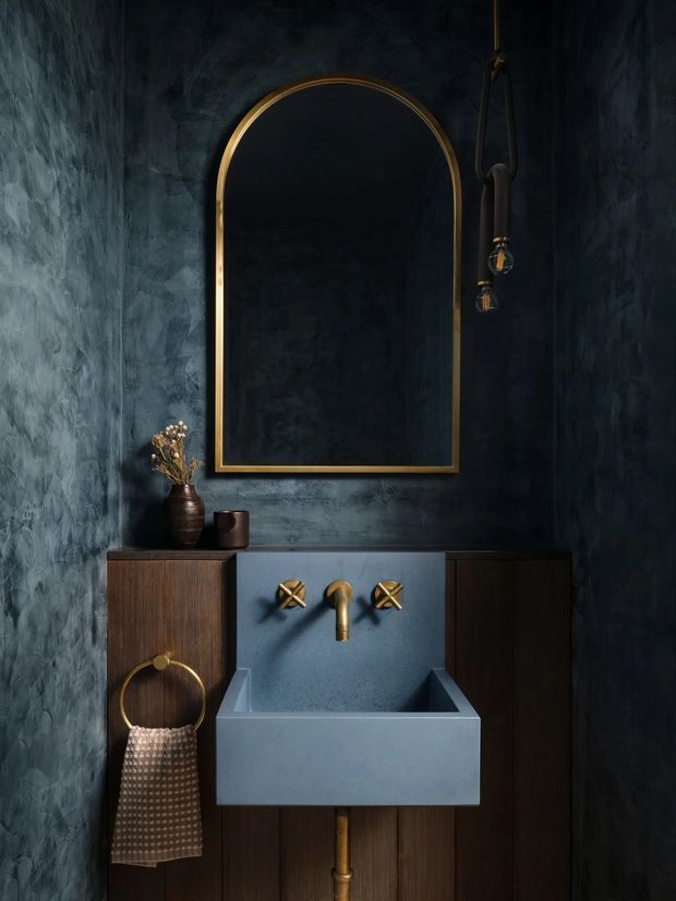 A moody, compact bathroom vanity vignette with a deep blue-grey textured wall, dark timber cabinetry, and a large arched mirror with a slim brass frame. The wall-mounted basin is blue-grey with exposed brass tapware, while a brass towel ring, dark vase, and hanging decorative light add refined detailing.
