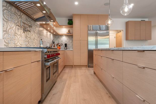 A warm, contemporary kitchen with extensive light timber cabinetry, a large central island, stainless-steel appliances, and a decorative patterned tile backsplash. The room features pale timber flooring, a dark stone or composite benchtop, open shelving, pendant globe lights, and a timber-slatted ceiling detail with integrated lighting.