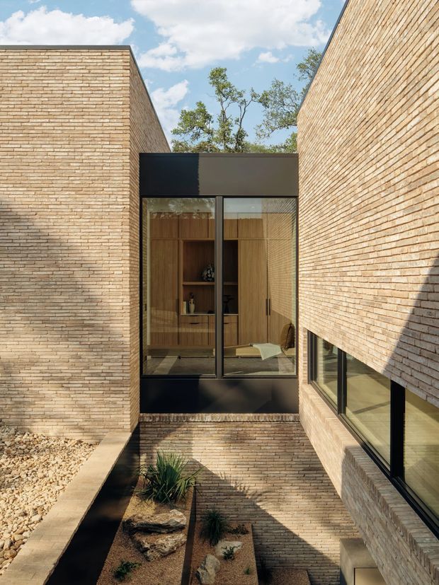 A contemporary brick-clad courtyard passage between two building wings. Tall pale brick walls frame black-framed floor-to-ceiling glazing and a projecting dark metal-framed connection. Through the glazing, warm timber built-in joinery is visible. A narrow landscaped sunken courtyard with gravel, rocks and drought-tolerant planting occupies the lower level, while mature trees and blue sky appear above.