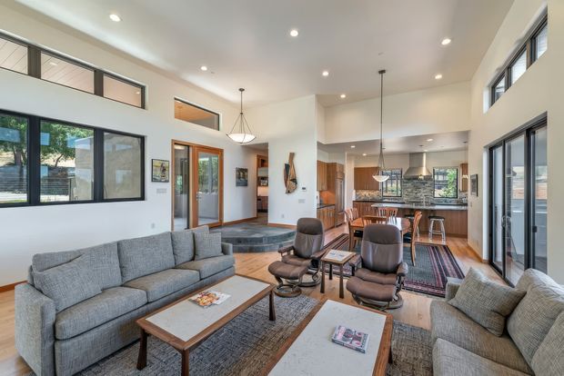 Open-plan contemporary living and dining area with a high, partially vaulted ceiling and abundant glazing. The room features grey upholstered sofas, two leather lounge chairs, timber-framed coffee tables, patterned area rugs, recessed lighting, pendant lights, and a dining zone leading into a modern kitchen with timber cabinetry and a tiled backsplash. Clerestory windows and large sliding doors create a bright, airy interior.