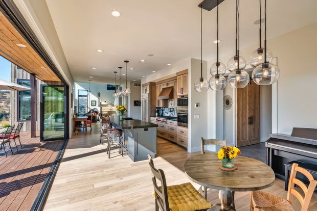 Bright open-plan kitchen and dining area with a long island, integrated timber cabinetry, stainless-steel appliances, pendant lighting, and a round timber dining table. Full-height glazed sliding doors open onto a timber deck, creating a strong indoor-outdoor connection. A living area is visible in the background.