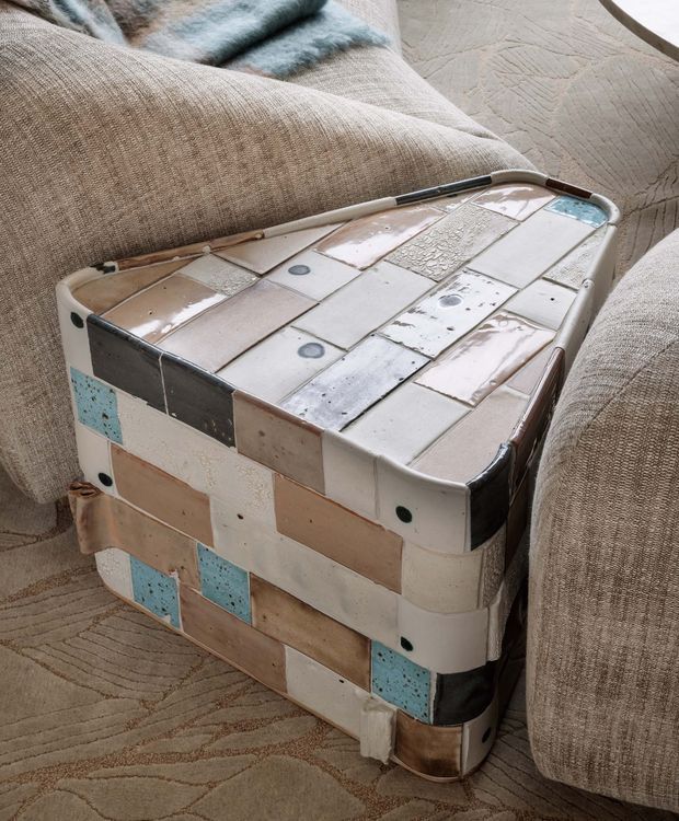 Close-up interior photograph of a vintage hard-sided suitcase or trunk repurposed as a small side table, covered in a patchwork of pale ceramic or tile-like panels with black, beige, blush, and turquoise accents. It sits between upholstered beige seating on a neutral patterned rug, with a soft blue throw visible in the background. The image emphasizes eclectic, tactile materials and a handcrafted decorative object.